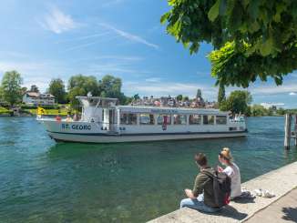 A small boat is sailing on the Rhine. Two people sit on the banks of the Rhine and look out over the river.