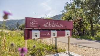 A purple-colored charging station stands in front of a parking lot. The station has three connections and a cabinet with various cables.