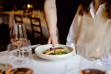 Ein Teller mit einem hübsch angerichteten Fischgericht wird auf einen weiss gedeckten Tisch gestellt. Auf dem Tisch stehen auch mehrere Weingläser mit Rosé.