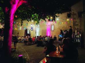 nordArt Stein am Rhein Festival benches in an illuminated courtyard. The benches are filled with people. A kleine Bühne has been set up. A musician is playing on it.