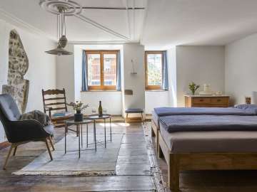 In a room with a wooden floor, there is a double bed, an armchair, and a wooden chair. The white plastered wall has an opening revealing the historic masonry.