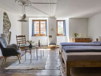 In a room with a wooden floor, there is a double bed, an armchair and a wooden chair. There is a hole in the white plastered wall where the historic wall is exposed.