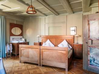 A double wooden bed with white bed linen. An old filigree parquet floor. The walls are beige and the wooden door has scenic paintings.
