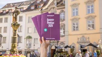 Eine Person hält das Genuss-Booklet in die Luft. Eine Broschüre mit Genusstipps und Gutscheinen zum Einlösen.