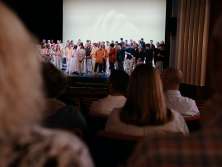 A theater stage with many artists. Spectators sit on the red seats in the auditorium.