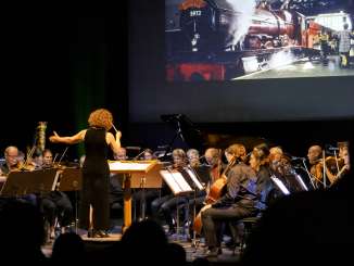 A conductor stands on stage with the orchestra. Behind them, on a screen, you can see a steam locomotive.
