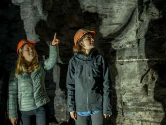 Three people are standing in a mountain tunnel. They are all wearing helmets. Someone is pointing at the ceiling with their index finger.