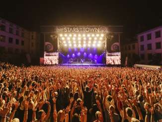 Stars in the city Many people with their arms in the air are at a concert with a band on a large stage.
