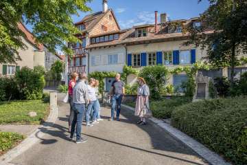 A woman in a traditional costume stands in front of a group and tells stories. The group is standing in an alley with houses with colorful facades.
