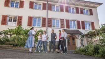 Guided tour of Hallau Five people stand in front of a house with red shutters. A guide in traditional costume tells village stories.