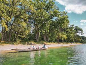 Three people and a dog are sitting in the Weidling, a long wooden boat. A woman is standing at the back, poking along the shore with its pebble beach and trees.