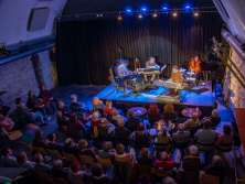 A kleine Bühne with four musicians. The full hall listens intently. The lighting is blue and red.