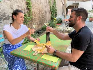 Two people are sitting at a table in a courtyard. They are eating a vegetable tarte flambée and toasting with a glass of white wine.