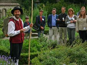 A person dressed in historical clothing and holding a wooden stick and a group of 7 people are standing in a herb garden. The group listens intently to the disguised person.