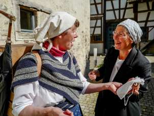 Two women dressed as shopkeepers chat with each other.