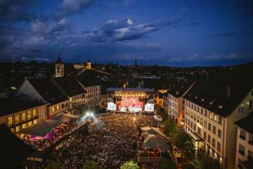 A square between houses in the old town. The square is filled with people. In front is a stage with the inscription "Stars in Town".