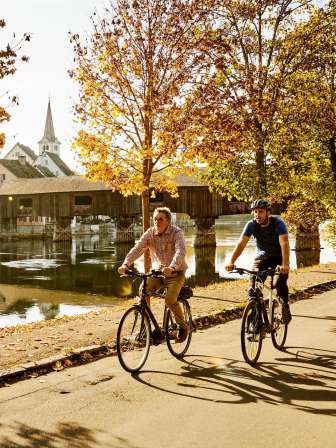 Two friends are cycling along the Rhine. There are golden trees on the banks and a wooden bridge over the Rhine.