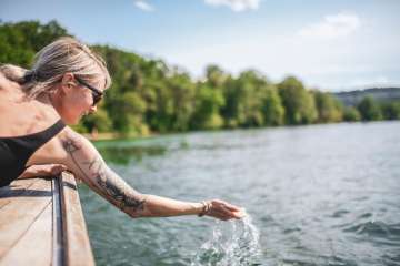 A woman is wearing a black swimsuit and sunglasses. She is lying on a wooden boat and holding one hand in the water.