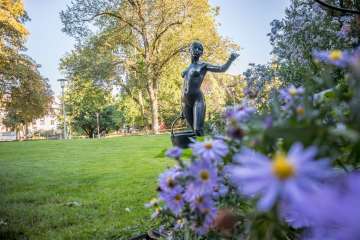 A statue of a woman carrying a basket in a park. In the foreground there are blooming flowers and in the background there are trees.