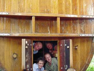 A huge old wine barrel with a door built into it. Four adults and a baby look out of the door from inside the barrel.