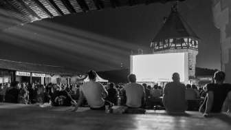 Many people are sitting on the ground or on benches, watching a film on a large screen in front of the Munot tower.