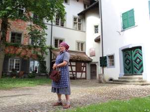 A woman is wearing a purple-blue apron and robe and a purple headscarf. She is standing in an inner courtyard.