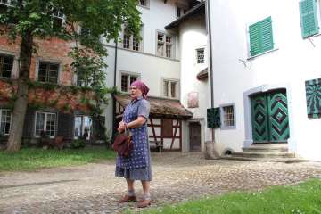 A woman wears a violet-blue apron and gown, along with a purple headscarf. She stands in a courtyard.