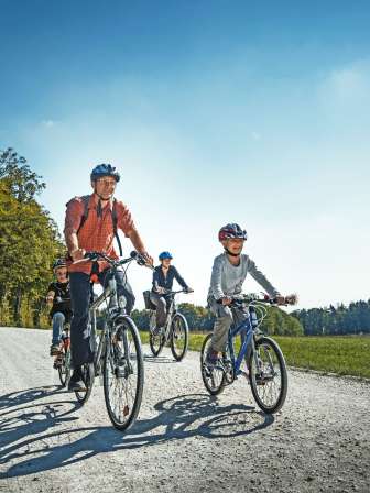 A family is cycling along a country lane. They are surrounded by meadows, fields and a forest.