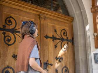 Schallhausen Music Trail A woman is wearing black "over the ears headphones". She is standing in front of the wooden door of a church. She is wearing a brown woolen sweater around her shoulders.