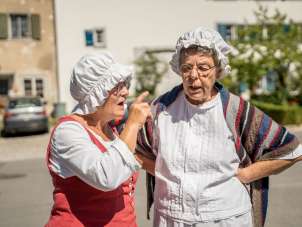 Two women in robes and white bonnets are chatting with each other.