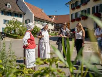 Two ladies in disguise tell a story. The rest of the people listen and laugh. There are old houses in the background.