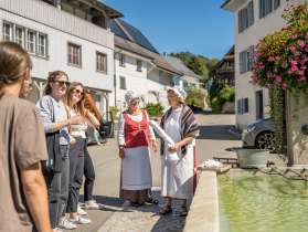 At a village fountain, two women dressed as 'Wöschwiiber' (washerwomen) with traditional garments and bonnets are standing. A group listens to them telling stories.