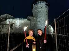 A man and a woman are standing with a burning torch each in front of Munot Fortress. She is also holding a lantern with a candle.