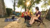 Two friends are sitting on lounge chairs and clinking their beers together. Their tent is set up behind them.