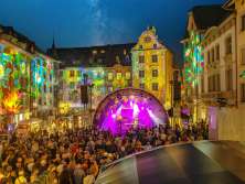 A square between houses. The facades are colorfully illuminated. In the middle is a stage with musicians. The square is filled with people.