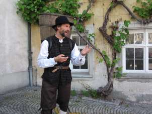 A person with historical clothing and a large wooden rucksack (a pannier). He is standing in front of a house facade where a vine is growing.