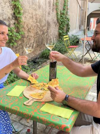 Two people are sitting at a table in a courtyard. They are eating a vegetable tarte flambée and toasting with a glass of white wine.