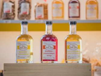 Drei verschiedene Sirupe mit den Geschmäcker Holunder, Himbeer-Johannisbeer und Löwenzahlblüte mit Orange stehen auf einer Holzkiste.