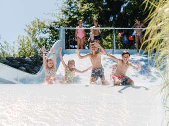 Four children slide down a wide slide together in an outdoor pool.