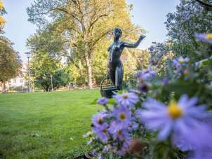 A statue of a woman carrying a basket in a park. In the foreground there are blooming flowers and in the background there are trees.