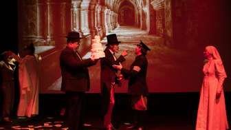 Sechs verkleidete Theaterspielende. Ein grossen Bild von einem Kreuzgang im Hintergrund. Eine Nonne, ein Polizist, ein Herr mit Zilinder, ein Herr mit einer Torte in der Hand.