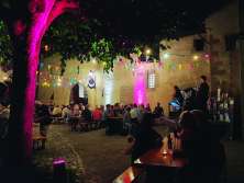 Festival benches in an illuminated courtyard. The benches are filled with people. A kleine Bühne has been set up. A musician is playing on it.