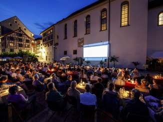 Many people are sitting in a square in front of a church, watching a movie that will be projected onto the church's facade,