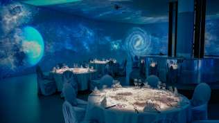 A galactic world is projected onto the walls, ceiling, and floor. Festively set tables stand in the room.