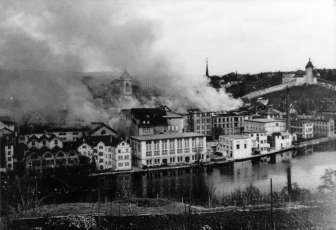 An old black and white picture of a city. Dark clouds are rising above it.