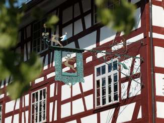 Blick auf eine eiserne Tafel mit einem goldenen Hirsch, die an einem Riegelhaus mit rötlichen Blaken befestigt ist.