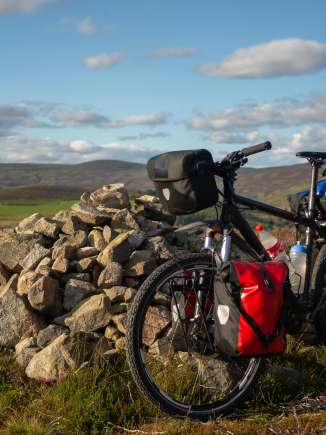 A bike with luggage A fully loaded touring bike stands at the side of the path, with a slightly hilly landscape stretching out behind it.