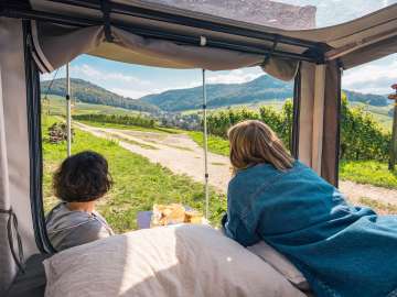 A woman is lying in a tent set up on a cargo bike, looking out at the vineyard landscape. A person is sitting in front of the tent, eating breakfast.