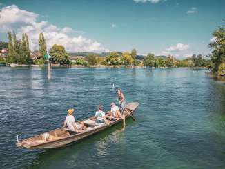 Three people and a dog are sitting in the Weidling. The wooden boat drifts leisurely down the Rhine. A woman is standing, holding an oar and steering the boat.