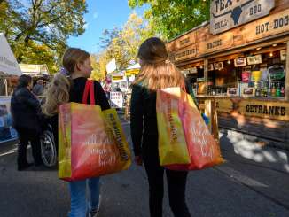 Zwei Mädchen mit je einer grossen Einkaufstasche, wo Herbstmesse darauf steht, spazieren zwischen Ständen hindurch.